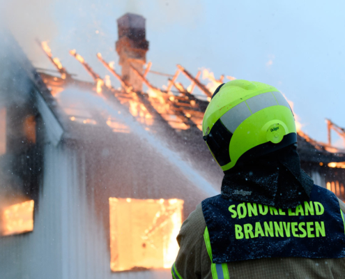Brannmann fra Søndre Land Brannvesen slokker pipebrann. Nesten hele taket er tatt av flammene